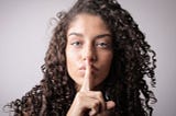 Woman with brown curly hair doing the be silent sign by covering her lips with a finger.