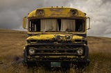 Old, rusted bus, with curtains hanging in the front windows, sitting in a field.