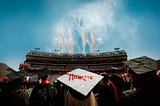Fireworks in the shape of Ns erupt behind Memorial Stadium as graduates watch. A graduate whose mortarboard reads “Huskers” is centered in the foreground.