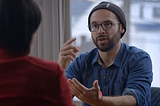 Photo of Aza Raskin being interviewed. He’s wearing glasses, facial hair, a beanie, and a button-down denim shirt as he gestures toward an interviewer at the edge of the frame.