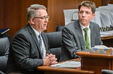 Two men in suits sit behind a wooden table. One of them speaks into a microphone as the other looks on.