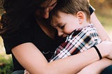 Mother holding a young child on her lap and hugging him close in a reassuring manner. The mother and child’s face are cast down. The mother is wearing a dark shirt and the son is wearing a plaid patterned shirt.