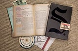 Descrição da imagem: a foto mostra o conjunto da publicação. Existe um livro aberto, com páginas amareladas e anotações à caneta feita nas margens, guardanapos desenhados, uma bússola, um pedaço de uma carta, um pedaço de uma foto e uma capa, preta com o símbolo de um S. e um macaco, onde o livro é guardado.