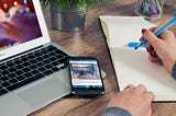 A writer on her desk with a laptop, a mobile phone, a notepad, and a pen