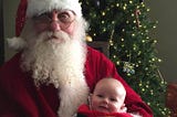 Bearded man with round gold frames sits in front of a Christmas tree in a Santa suit with a baby on his lap.