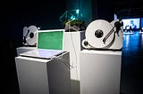 A laptop, two vinyl players and the moss installation each of them placed on a white pillar in an exhibition space.