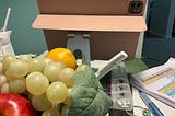 A close-up view of a table with various items, including an iPad in a pink case on a stand, a pile of green grapes, a red apple, an orange, a white cloth napkin with a straw, a white pen, a daily pill organizer, and some papers with highlighted text. The background shows a green wall and part of a doorway.