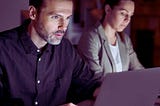 Zdjęcie przedstawia osoby pracujące przy laptopach w nocy. Mężczyzna ubrany w ciemną koszulę, ma skupiony wyraz twarzy i patrzy intensywnie w ekran, co sugeruje pełne zaangażowanie w wykonywane zadanie. W tle widać kobietę w jasnej marynarce, również pochyloną nad laptopem, co wskazuje na jej koncentrację i determinację. Obraz oddaje istotę wyzwania, jakim jest praca w nocy osób niepełnosprawnych — skupienie, determinację oraz wytrwałość w dążeniu do celu pomimo zmęczenia.