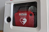 A red automated external defibrillator behind a glass window in a wall-mounted case.