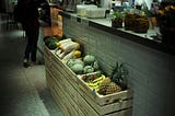 A row of wooden crates filled with fruit — bananas, pineapples, melons, and papayas — rests against a tiled wall under soft lighting. A person stands in the background near a counter, slightly out of focus. The atmosphere is quiet, casual, and intimate, evoking a moment paused in motion.