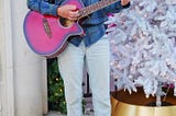 Photo of author as she strums her guitar and appears to be singing. She wears a light grey hat, purple glasses, light jeans, yellow Vans sneakers and a blue plaid shirt. There is a white Christmas tree behind her and faux pine with lights.