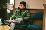 A young boy sits alone on a folding chair with his arms crossed and legs tucked in, wearing a green jacket and jeans. His expression looks distant and serious, as though he’s deep in thought or waiting.