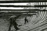 Two men building a wooden ship at an old shipyard.