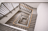 Overhead view looking down a modern interior stairwell with clean lines, neutral tones, and evenly spaced landings.