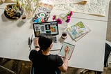 A woman sits at a table with her computer and sketch pad creating art.