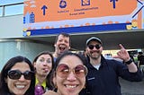 Five people smile for a selfie outdoors in front of a large orange “KubeCon + CloudNativeCon Europe 2026” event banner decorated with stylized buildings, tulips, a bicycle, and a bridge.