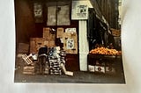 An a thin person in tattered clothes pokes through boxes of food in a New York City alley. On the street is a large display of fresh oranges.