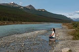 A woman with long dark hair sits on a rock in meditation at the edge of a river with mountain peaks on the other side.