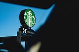 Image shows iconic green and white Starbucks sign reading “Drive Thru” against a bright blue sky background, viewed through the front windshield of a car. Foreground shows blurred, dark silhouette of car’s interior, including seat, visor, and steering wheel.