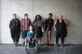 Six disabled people of colour smile and pose in front of a concrete wall.
