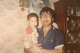 Mr. Lee holding baby Lisa in the living room of their home, both looking at the camera, dated May 4th, 1985.