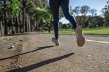 Legs and feet of an athlete running down a road in a green park surrounded by trees.