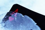 The left top corner of an Ace of Hearts card rises out of the ice. The card is black with a red A above a red heart. The ice is pale blue.