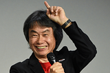 Shigeru Miyamoto on stage, pointing to the sky with a microphone in his hand.