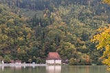 A building with red roof on the river bank, with autumn trees above.