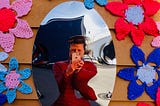 Nina Elliott takes a photo of herself in a distorted mirror surrounded by colourful crocheted flowers hung on an outside wall.