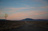 A desert sunset with subdued reds and yellows above a dirt track heading away from a stop sign visible only from behind.