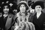 Black-and-white photo of four young women in hats and coats, including suffrage & labor activist “Fola” La Follette and social reformer and missionary Rose Livingston, in a 1913 New York City garment strike.