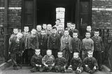 A black and white photo of about two dozen pre-adolescent boys in uniform jackets standing or sitting outside a brick building with what appear to be metal doors