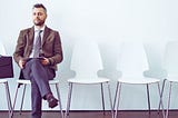 Mężczyzna w eleganckim garniturze siedzi na krześle w poczekalni przed rozmową o pracę, trzymając teczkę z dokumentami — przygotowuje się na pytania na rozmowie kwalifikacyjnej.