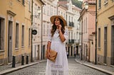 A woman in a white flowy dress and straw hat standing on a cobblestone street in a European city, showcasing a chic Europe outfit for summer travel.