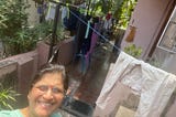 A woman wearing a red dot on her forehead (Southeast Asian Indian) smiles at the camera. There are clotheslines behind her where clothes dry. There are bright green plants beside the lady and behind the clotheslines. It is a bright, sunny day in India.