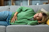 Mature woman alone at home sick, holding hands to her stomach, lying on a sofa.