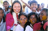 A smiling woman bedecked with a white and red flower garland surrounded by happy children.