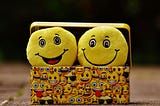 a yellow box with pictures of yellow emoticons all over them. Two plush emoticons smiling are under the open lid of the emoticon