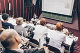 A lecture theatre with people sat in the audience and a presenter speaking.