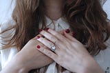 A woman with long brown hair folds her hands across her chest. She wears a silver ring with a cut-out of a heart.