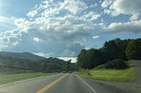 A highway in Vermont, lined with trees, and white clouds and mountains in the distance against a blue sky.