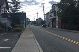 A long road two-lane road with a double yellow stripe down the middle a sidewalk to the left and buildings on either side. No traffic.