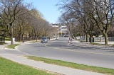 City street in daytime with a treelined streets and a car in the distance.