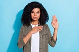 A black woman with one hand on chest the other raised taking a pledge in from of a blue background.