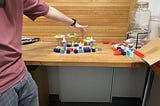 A person with their finger atop a model made from various shapes — cubes, cones, cylinders, all on a wooden table.