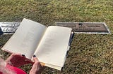 An open book is held in front of a gravestone. Long white hair and hands of the person reading are visible at the corner of the image.