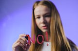 A girl writing no on the screen with a pink pen. She is lit with purple background.