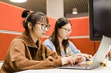 Two students sit and work on their laptops inside a booth in a library on campus. The booth has orange walls and grey seats.