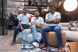 Three middle-aged south asian men sitting casually on brown and cream office furniture, dressed in jeans and white t-shirts with the Hola Health logo.